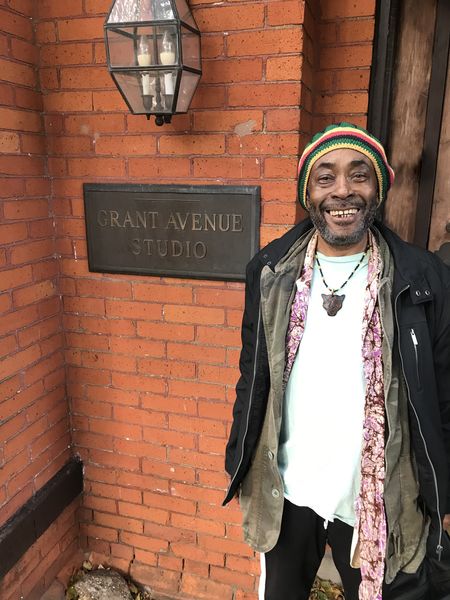 Klyde Broox author photo. A smiling man wearing a colorful knit hat, layered clothing, and a necklace stands outside a brick building beside a sign that reads “Grant Avenue Studio.” A lantern-style light fixture is mounted above the sign. Klyde Broox author photo. A smiling man wearing a colorful knit hat, layered clothing, and a necklace stands outside a brick building beside a sign that reads “Grant Avenue Studio.” A lantern-style light fixture is mounted above the sign.