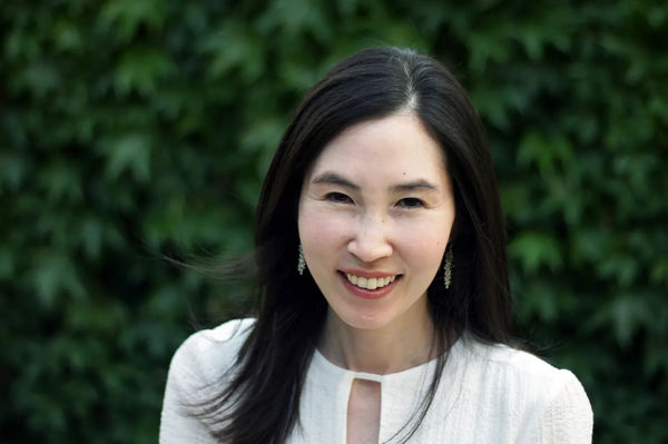 Leslie Shimotakahara author photo. A woman with long dark hair smiling at the camera, wearing a white blouse and dangling earrings, standing outdoors in front of a leafy green background.