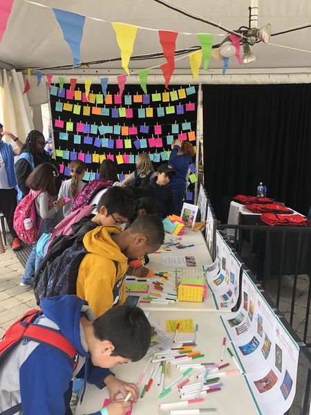 A number of young people writing letters to authors on a table with Lindsay Zier-Vogel in the background facilitating and rows of letters on string behind her.
