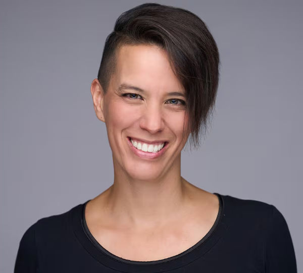 Author photo of Lindsay Diehl, showing a person with short dark hair styled longer on top and shaved on the sides, wearing a black shirt and smiling warmly at the camera against a gray background.