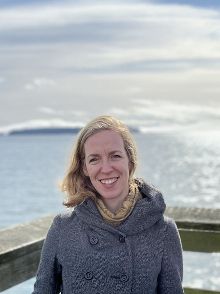 Author photo of Liz Johnston. A woman standing outdoors on a wooden pier with a calm body of water and distant land in the background. She is smiling, wearing a gray coat and a light scarf, with soft sunlight illuminating her face under a partly cloudy sky.