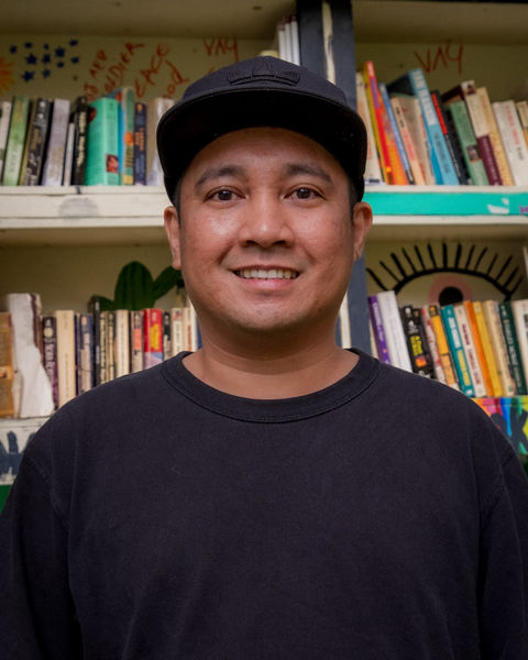 Marc Perez author photo. Man in baseball cap and t-shirt smiling and standing squarely to reader with bookshelves behind him in background.