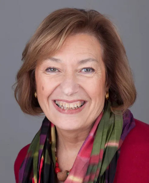 Professional headshot of Maria Coletta McLean against a neutral gray background. She has shoulder-length blonde hair with darker roots styled in soft waves, and wears gold hoop earrings. She is smiling warmly and broadly at the camera, wearing a red top with a colorful silk scarf in shades of green, pink, purple, and gold draped around her neck. Professional headshot of Maria Coletta McLean against a neutral gray background. She has shoulder-length blonde hair with darker roots styled in soft waves, and wears gold hoop earrings. She is smiling warmly and broadly at the camera, wearing a red top with a colorful silk scarf in shades of green, pink, purple, and gold draped around her neck.
