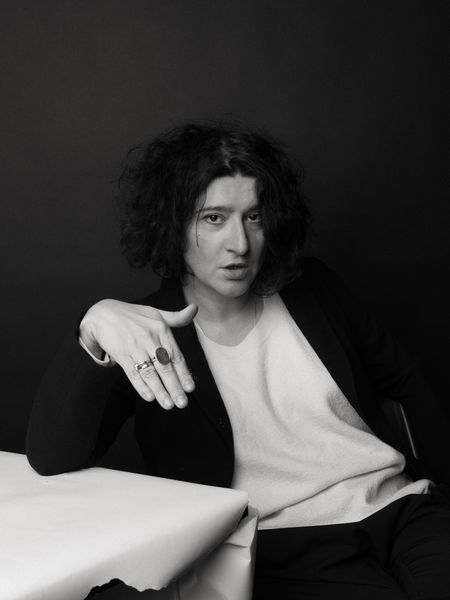 Maria Stepanova (Photo by Andrey Natotsinsky). Black-and-white portrait of a person with curly dark hair wearing a light sweater and dark blazer, seated at a table with one hand resting forward, showing several rings. The background is plain and dark, giving the image a dramatic, artistic tone.