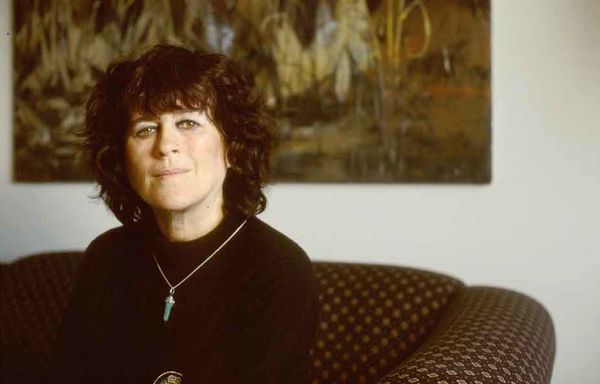 Marie-Claire Blais (Photo by Jill Glessing). A person with curly dark hair sits on a patterned sofa, wearing a black top and a necklace with a turquoise pendant. They are looking slightly toward the camera with a calm expression. A large abstract painting hangs on the wall behind them. Marie-Claire Blais (Photo by Jill Glessing). A person with curly dark hair sits on a patterned sofa, wearing a black top and a necklace with a turquoise pendant. They are looking slightly toward the camera with a calm expression. A large abstract painting hangs on the wall behind them.