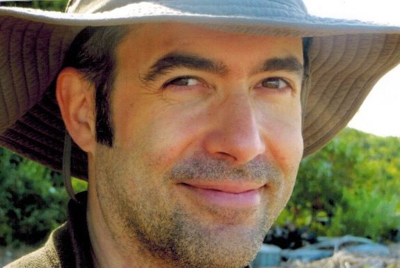 Author Mark Dickinson, outdoors and smiling at the camera. He wears a wide-brimmed sun hat and a dark shirt, with greenery and sunlight in the background.