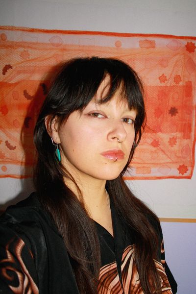 Portrait of illustrator Maya McKibbin with long dark hair and bangs, wearing a black patterned shirt and a turquoise earring, posed against a warm orange textile backdrop.