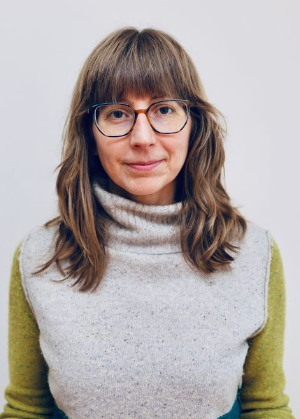 Author Melanie Dennis Unrau, photographed by Jason Unrau. She is shown from the shoulders up, wearing glasses and a light grey turtleneck sweater with green sleeves. She has medium-length brown hair and is standing against a plain light background, looking softly at the camera. Author Melanie Dennis Unrau, photographed by Jason Unrau. She is shown from the shoulders up, wearing glasses and a light grey turtleneck sweater with green sleeves. She has medium-length brown hair and is standing against a plain light background, looking softly at the camera.