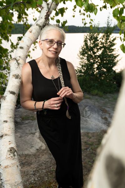 Melanie Marttila (Photo by Gerry Kingsley) - Image of woman with black dress and grey hair smiling and leaning against a birch tree.