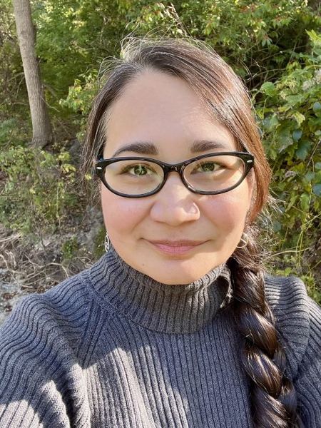 Author photo of Melissa Powless Day, a woman with long dark hair in a braid, wearing black glasses and a gray ribbed turtleneck sweater, smiling softly at the camera while standing outdoors with green foliage and trees in the background.
