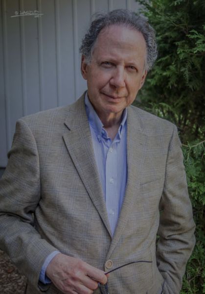Author photo of Michael Carin. Portrait of an older man with short gray hair, standing outdoors beside a green hedge and a light-colored wall. He is wearing a beige plaid blazer over a light blue collared shirt and looks directly at the camera with a calm, confident expression, one hand resting near his jacket button. Author photo of Michael Carin. Portrait of an older man with short gray hair, standing outdoors beside a green hedge and a light-colored wall. He is wearing a beige plaid blazer over a light blue collared shirt and looks directly at the camera with a calm, confident expression, one hand resting near his jacket button.