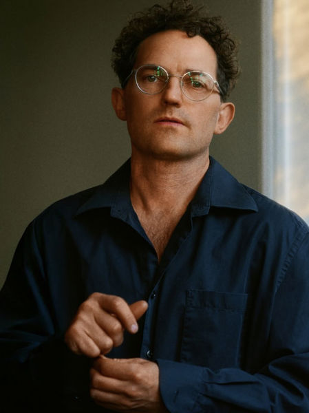 Author photo of Michael Eddy. He stands in three-quarter view against a softly blurred interior with warm daylight streaming from the right. He wears a navy blue button-up shirt with chest pockets and vintage-style wire-frame glasses with a subtle green tint. His dark curly hair frames his face, and he gazes directly at the camera with a calm, thoughtful expression. His hands are positioned at mid-torso, one resting over the other in a relaxed gesture. The lighting is warm and natural, casting soft shadows and highlighting the texture of the fabric and the contours of his face. The background suggests an architectural space with muted olive and cream tones, keeping the focus on the subject. The overall mood is intimate, grounded, and quietly confident.