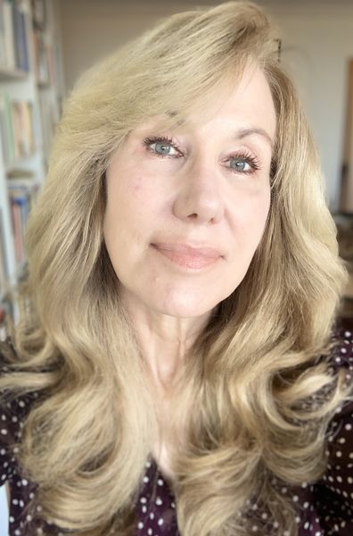 Auhtor photo of Moira Welsh. A woman with long, wavy blonde hair and light eyes, wearing a dark patterned top, poses for a portrait indoors with bookshelves softly blurred in the background. Auhtor photo of Moira Welsh. A woman with long, wavy blonde hair and light eyes, wearing a dark patterned top, poses for a portrait indoors with bookshelves softly blurred in the background.