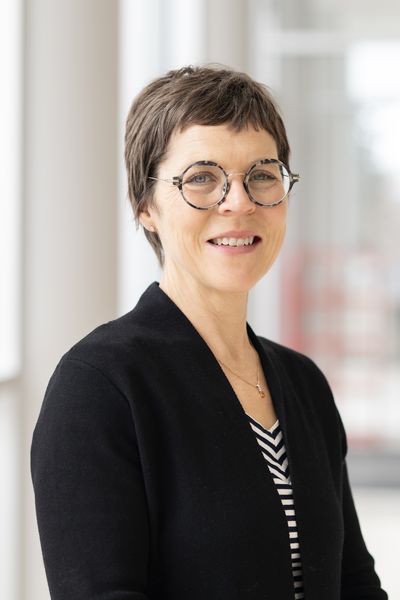 Author Nadia Bozak smiling in a portrait against a softly blurred indoor background, wearing round glasses, a black jacket, and a striped top. Author Nadia Bozak smiling in a portrait against a softly blurred indoor background, wearing round glasses, a black jacket, and a striped top.