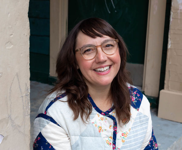 Author photo of Naseem Hrab. She is smiling warmly at the camera, wearing clear-framed glasses and a colorful patchwork sweater featuring white textured fabric, navy blue sections, and floral embroidered details in yellow and pink. She has shoulder-length brown wavy hair with side-swept bangs. The photo is taken outdoors against a cream-colored brick wall with a window visible in the background.