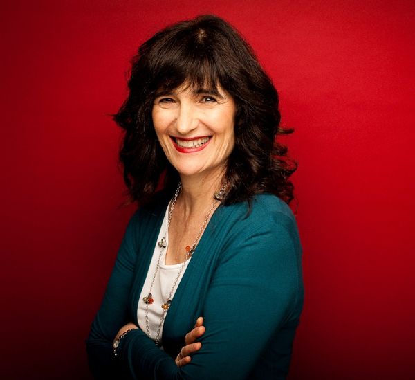 Author photo of Nora Gold. A woman with shoulder-length dark brown wavy hair with bangs smiles warmly at the camera against a vibrant red background. She wears a teal cardigan over a white top, accessorized with a long silver necklace featuring colorful pendant charms. Her arms are crossed in a relaxed, confident pose, and she wears bracelets on her wrist. The professional portrait captures her approachable and engaging presence.