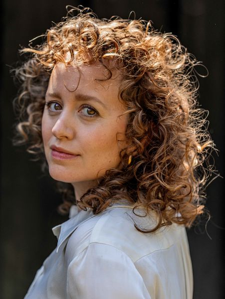 Author Olga Ravn, with curly light brown hair wearing a light-colored shirt, looking over their shoulder toward the camera with a calm expression, against a dark blurred background. Author Olga Ravn, with curly light brown hair wearing a light-colored shirt, looking over their shoulder toward the camera with a calm expression, against a dark blurred background.
