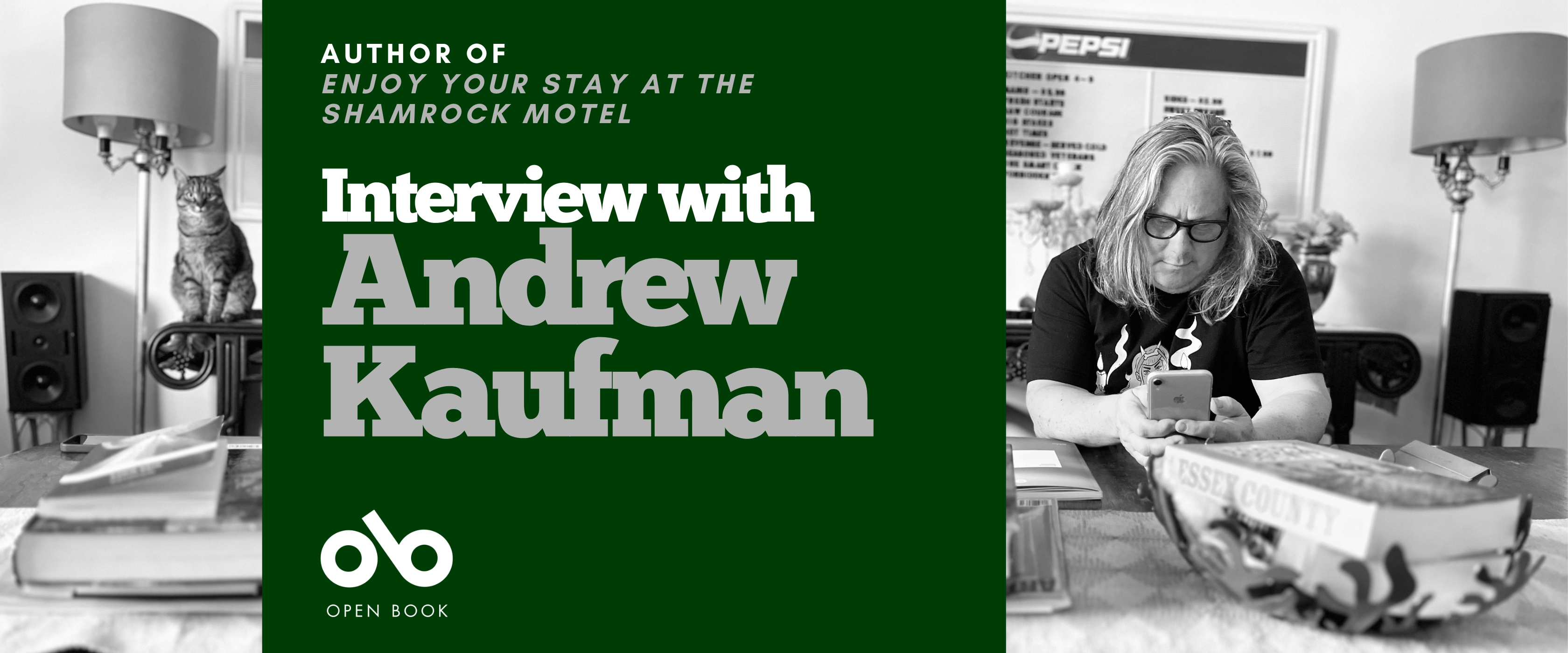 Interview with Andrew Kaufman, author of Enjoy Your Stay at the Shamrock Motel. Background image of author at his large, busy desk in a crowded room with various household trappings and a cat sitting hilariously on the edge of a chair. Author a man with long, grey hair and a black t-shirt, and glasses, looking down at his phone with elbows on the same table. Solid green area to centre left of banner with text and Open Book logo overlaid.