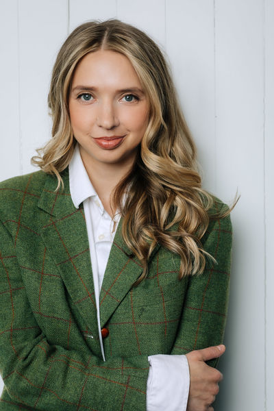 Author photo of Pippa Mackie. She has long, wavy blonde hair and is wearing a green tweed blazer with an orange windowpane pattern over a white collared shirt. She is smiling warmly at the camera against a white paneled background.