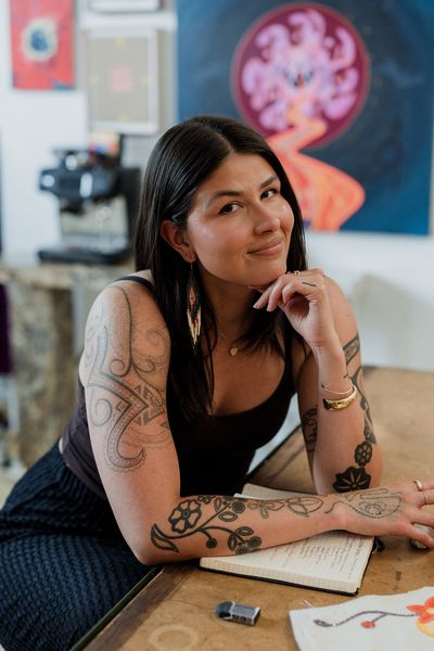 Author Quill Christie-Peters, photographed by Tenille Campbell, sits at a wooden table with a notebook and artwork in front of her. She rests her chin lightly on her hand and smiles at the camera. Her tattooed arms and long beaded earrings are visible, and behind her hangs a vibrant painting featuring swirling pink and orange tones. The setting feels creative and warm, capturing an artist at work. Author Quill Christie-Peters, photographed by Tenille Campbell, sits at a wooden table with a notebook and artwork in front of her. She rests her chin lightly on her hand and smiles at the camera. Her tattooed arms and long beaded earrings are visible, and behind her hangs a vibrant painting featuring swirling pink and orange tones. The setting feels creative and warm, capturing an artist at work.