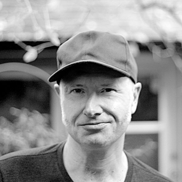 Author photo of Ray Robertson. Black-and-white portrait of a person wearing a dark baseball cap and a T-shirt, standing outdoors in front of a blurred background with a building and foliage. The person is looking at the camera with a slight smile. Author photo of Ray Robertson. Black-and-white portrait of a person wearing a dark baseball cap and a T-shirt, standing outdoors in front of a blurred background with a building and foliage. The person is looking at the camera with a slight smile.