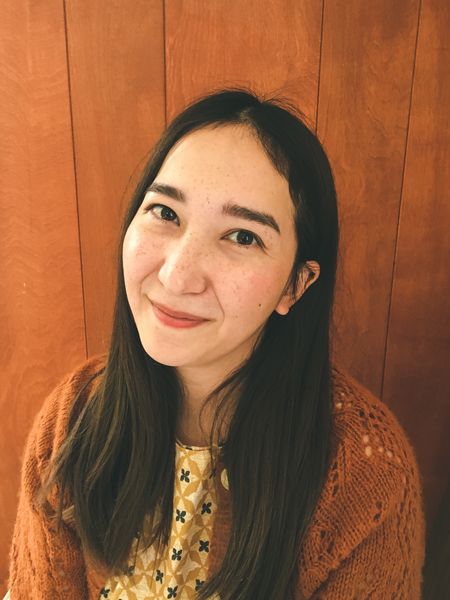 Risa Hugo, woman with long straight brown hair smiling softly at the camera. She is wearing a mustard-yellow patterned top and a rust-colored knit cardigan, sitting in front of a warm wooden wall. The lighting is soft and natural, giving the portrait a cozy, inviting feel.