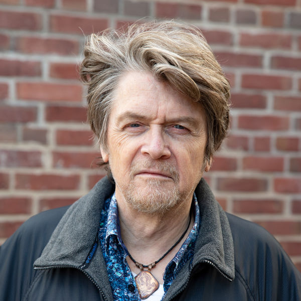 Author Robert Priest standing in front of a brick wall, wearing a patterned blue shirt, a dark jacket, and a pendant necklace, looking directly at the camera.