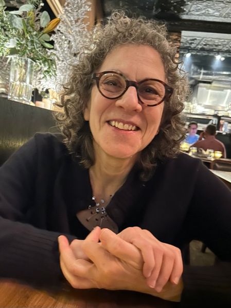 Author photo of Ronna Bloom, a person with short curly gray hair and round glasses smiling while seated at a restaurant table. They are wearing a black sweater, and the background shows a warmly lit dining area with other patrons and decorative plants.