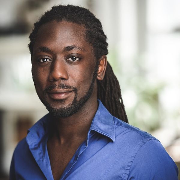 Portrait of author Ryad Assani-Razaki, wearing a blue collared shirt, looking at the camera with a calm expression. His long hair is styled in dreadlocks, and the background is softly blurred.