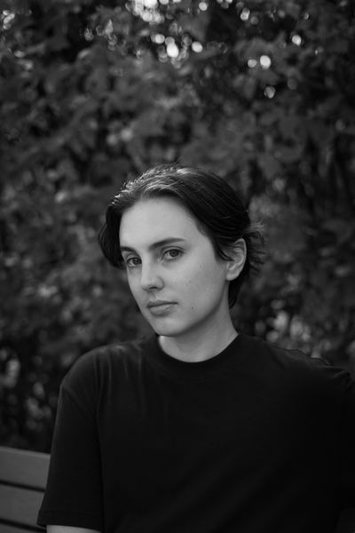 Black-and-white author portrait of Sam K MacKinnon. The subject sits facing the camera with a calm, direct gaze, wearing a simple black crew-neck shirt. Their dark hair is pulled back loosely, with soft natural light illuminating their face and creating gentle contrast. The background is softly blurred foliage with dappled light filtering through leaves, giving the image an organic, contemplative quality. The composition is centered and intimate, emphasizing the author's thoughtful presence. Photo credit: Bryce Hoye.