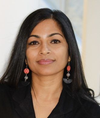 Author photo of Saraswoti Lamichhane. A woman with long, straight black hair looks directly at the camera with a warm, gentle smile. She wears distinctive statement earrings featuring coral-pink circular elements and black tassels. She's dressed in a black top and a delicate gold necklace. The background is softly blurred in neutral tones. The portrait conveys an approachable and confident presence.