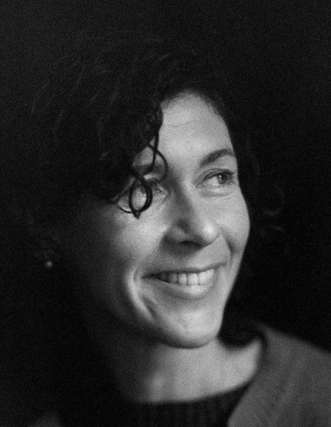 Author photo of Sasha Dugdale, Translator of The Disappearing Act. Black-and-white portrait of a person with short curly hair smiling softly and looking to the side. They are wearing a sweater over a dark top, and the background is dark and softly blurred.