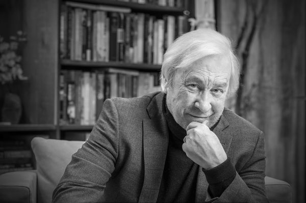 Black and white portrait of Scott McIntyre seated indoors, wearing a blazer and turtleneck. He rests his chin on his hand and looks at the camera with a slight smile. A bookshelf filled with books is visible in the background. Photo by Vasgen Degirmentas.