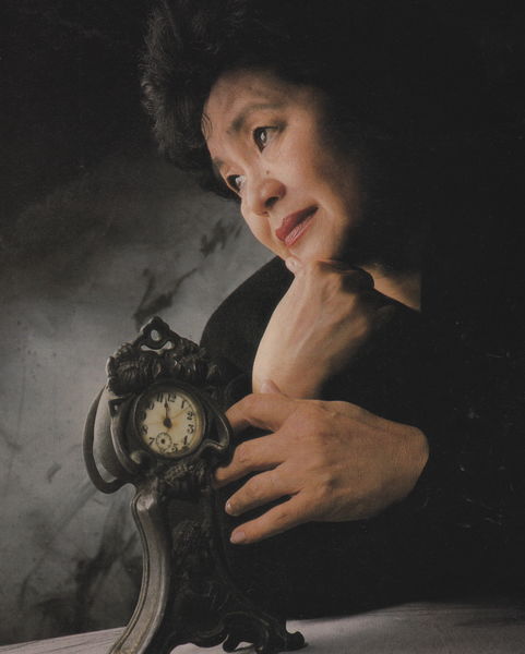 Portrait of Setsuko Thurlow, shown in soft lighting with a contemplative expression. She is resting her chin on one hand while gently holding an ornate, antique-style clock with the other. She wears a dark outfit against a shadowy background, evoking a reflective and somber mood. The clock’s hands are stopped at a specific time, symbolizing a poignant moment in history.