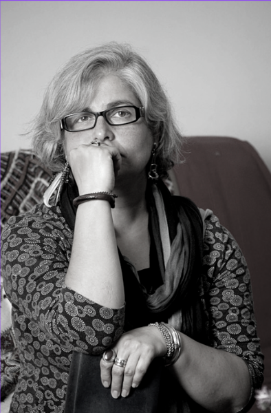 Black-and-white portrait of Shahrzad Arshadi, seated indoors with her chin resting on her hand. She wears glasses, a patterned top, bracelets, and a scarf, and looks thoughtfully off-camera.