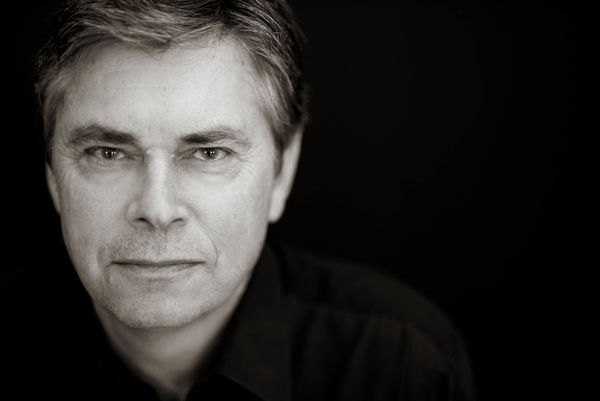 Black-and-white author portrait of a man shown from the shoulders up against a dark background. He has short, light hair and a calm, direct expression, wearing a dark collared shirt. The photo is sharply lit, emphasizing his thoughtful gaze and the texture of his features. Photo by Peg McCarthy.