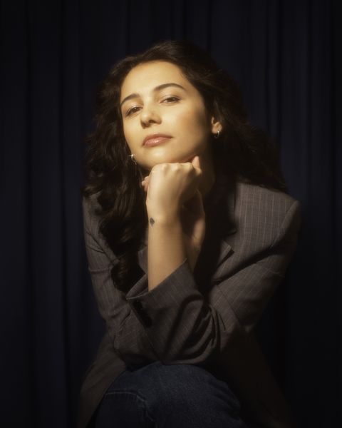 Author photo of Sierra Marilyn Riley (Photo by Tessla Stuckey). A woman with long, wavy dark hair poses against a dark curtain backdrop. She rests her chin on her hand and looks confidently at the camera. She is wearing a gray pinstripe blazer and jeans, with soft, warm lighting giving the portrait a vintage, cinematic feel. Author photo of Sierra Marilyn Riley (Photo by Tessla Stuckey). A woman with long, wavy dark hair poses against a dark curtain backdrop. She rests her chin on her hand and looks confidently at the camera. She is wearing a gray pinstripe blazer and jeans, with soft, warm lighting giving the portrait a vintage, cinematic feel.