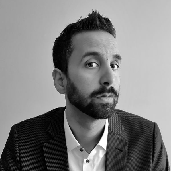 Sunny Dhillon (Photo by Sunny Dhillon). Black-and-white portrait of a man with short dark hair and a trimmed beard, wearing a pinstriped suit jacket over a white collared shirt. He looks directly at the camera with a neutral expression against a plain light background.