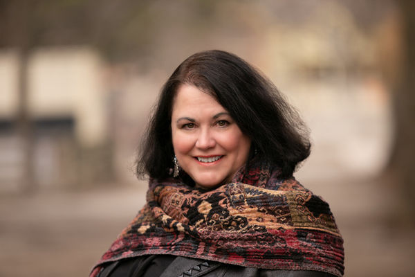 Author photo of Susan Currie, woman with shoulder-length dark brown hair smiles warmly at the camera. She is wearing a patterned shawl in shades of burgundy, brown, and gold over a dark top. The background is softly blurred, showing an outdoor setting with muted trees and buildings, creating a calm, natural atmosphere. Author photo of Susan Currie, woman with shoulder-length dark brown hair smiles warmly at the camera. She is wearing a patterned shawl in shades of burgundy, brown, and gold over a dark top. The background is softly blurred, showing an outdoor setting with muted trees and buildings, creating a calm, natural atmosphere.
