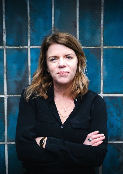 Author Susanna Crossman standing against a blue-tiled wall, wearing a black shirt with layered necklaces, arms crossed, and looking directly at the camera.