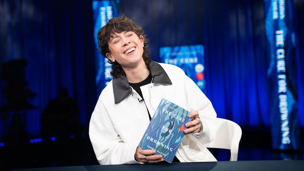 Tegan Quin wins Canada Reads 2026, championing Loghan Paylor's novel, The Cure for Drowning. (Joanna Roselli/CBC). A person with short dark hair laughing joyfully while holding 'The Cure for Drowning' by Loghan Paylor, wearing a white jacket with black collar, seated against a blue-lit stage backdrop with Canada Reads branding visible.