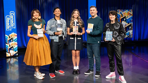 The 2026 Canada Reads contenders from left to right, Elle-Máijá Tailfeathers, Josh Dela Cruz, Morgann Book, Steve (Dangle) Glynn and Tegan Quin. (Joanna Roselli/CBC). Five people stand on a stage holding books, smiling at the camera. Behind them are blue curtains and signs that read “Canada Reads.” The group is dressed in a mix of casual and stylish outfits, and each person is holding a different book featured in the competition.