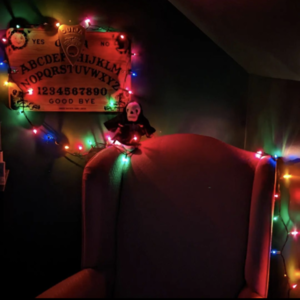 A pink wingback chair underneath a wall-mounted Ouija board, lit by Christmas lights.
