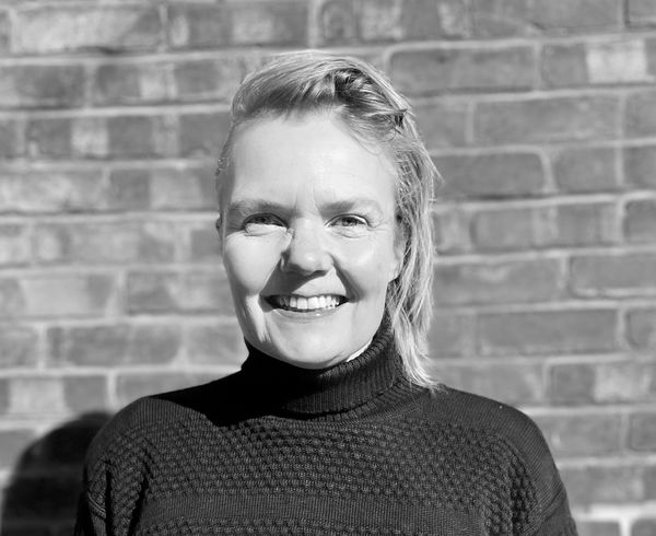Tine Modeweg-Hansen (Photo by Allie Brown). Black-and-white portrait of a smiling woman standing in front of a brick wall. She has short, light-colored hair brushed to the side and is wearing a textured, high-neck sweater. The sunlight casts soft shadows, highlighting her face and creating a warm, natural feel. Tine Modeweg-Hansen (Photo by Allie Brown). Black-and-white portrait of a smiling woman standing in front of a brick wall. She has short, light-colored hair brushed to the side and is wearing a textured, high-neck sweater. The sunlight casts soft shadows, highlighting her face and creating a warm, natural feel.