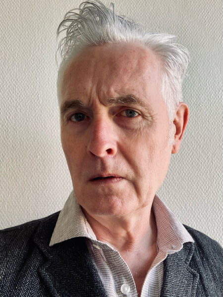 Photo of translator Martin Aitken, man with short gray hair wearing a collared shirt and dark blazer, looking directly at the camera with a neutral expression against a light textured wall. Photo of translator Martin Aitken, man with short gray hair wearing a collared shirt and dark blazer, looking directly at the camera with a neutral expression against a light textured wall.