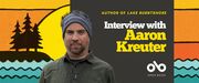 Interview with Aaron Kreuter, author of LAKE BURNTSHORE banner. Background image from cover of stylized, illustrated lakefront scene with undulating waves across bottom of image and dark silhouette of island with pines atop in to left, in front of a colourful setting sun with a sky in yellows and oranges. Solid brown section to centre right with text and Open Book logo overlaid. Image of author to centre left, man with a rugged headband and beard and an outdoorsy shirt, looking like he is going camping in the woods.