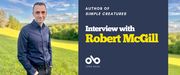 Interview with Robert McGill, author of Simple Creatures (Coach House Books) banner. Background image of sunny, rolling parkland with trees in distance. Author standing in foreground, smiling slightly with hair combed back nearly, wearing button up shirt and dark jeans with his hands in pockets. To centre right a solid blue section with text and Open Book logo overlaid.