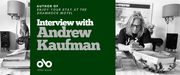 Interview with Andrew Kaufman, author of Enjoy Your Stay at the Shamrock Motel. Background image of author at his large, busy desk in a crowded room with various household trappings and a cat sitting hilariously on the edge of a chair. Author a man with long, grey hair and a black t-shirt, and glasses, looking down at his phone with elbows on the same table. Solid green area to centre left of banner with text and Open Book logo overlaid.