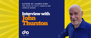 Interview with John Thurston banner. Background image of bold yellow and orange lines fanning out from a central focus point. Area of rich blue at centre-left of image with text and Open Book logo overlaid. Image to right of author, older man smiling and looking to reader, wearing a bold blue collared shirt with his arm crooked as if resting on something.