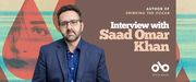 Interview with Saad Omar Khan, author of Drinking the Ocean banner. Background image from book cover of textured landscape blurred and tinted with red colours overlaid and shapes of teardrops in deeper reds, with images of a woman's face in some of them. Solid deep pink section to centre-right with text and Open Book logo overlaid. Image of author to centre-left, man in blue dress shirt and suit jacket with thick, short hair and a neat beard, wearing glasses and looking out pensively